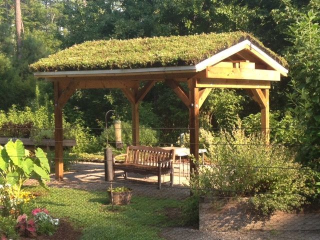 Wooden gazebo with green roof, bench, and plants in a garden setting.