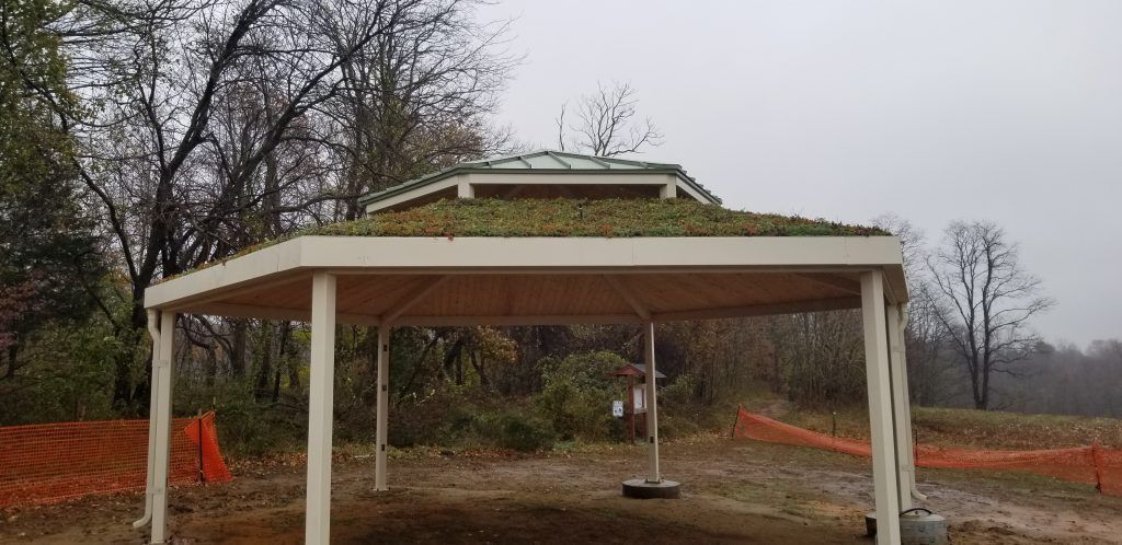 A white-pillared gazebo with a green roof, set in a wooded area with an overcast sky.