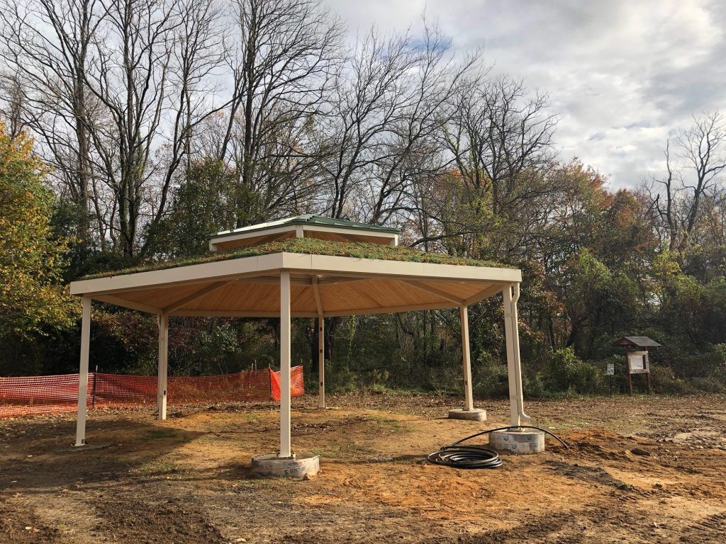 Pavilion with green roof in a dirt field, surrounded by trees and overcast sky.