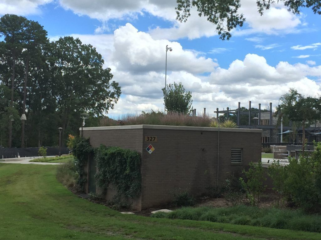 Brick building with a green roof, a grassy area, and a metal structure in the background under a cloudy sky.