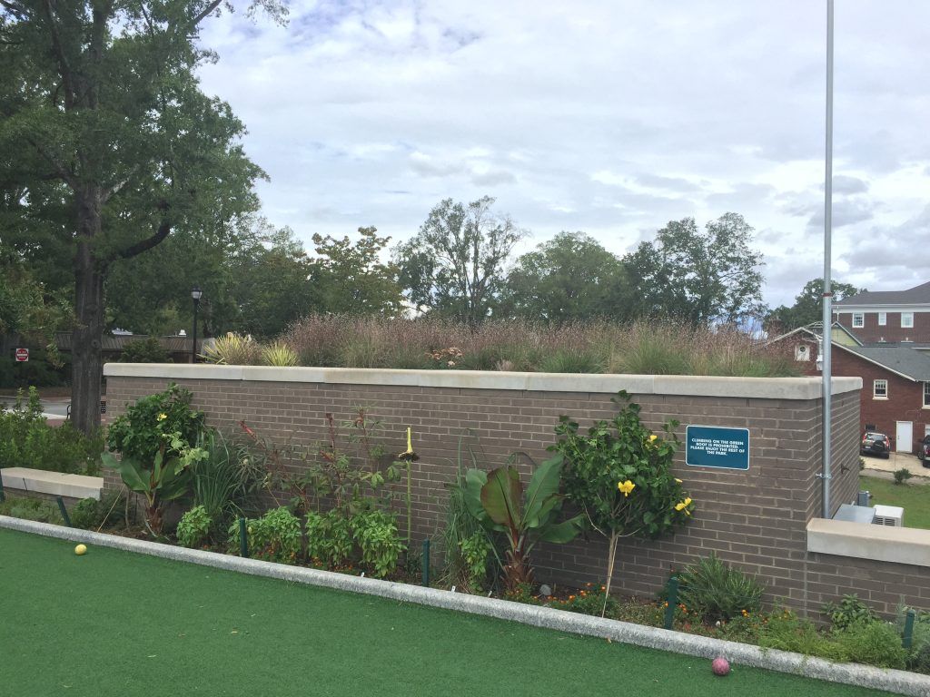 Brick wall with a green roof garden and plants below, green turf in foreground, cloudy sky.