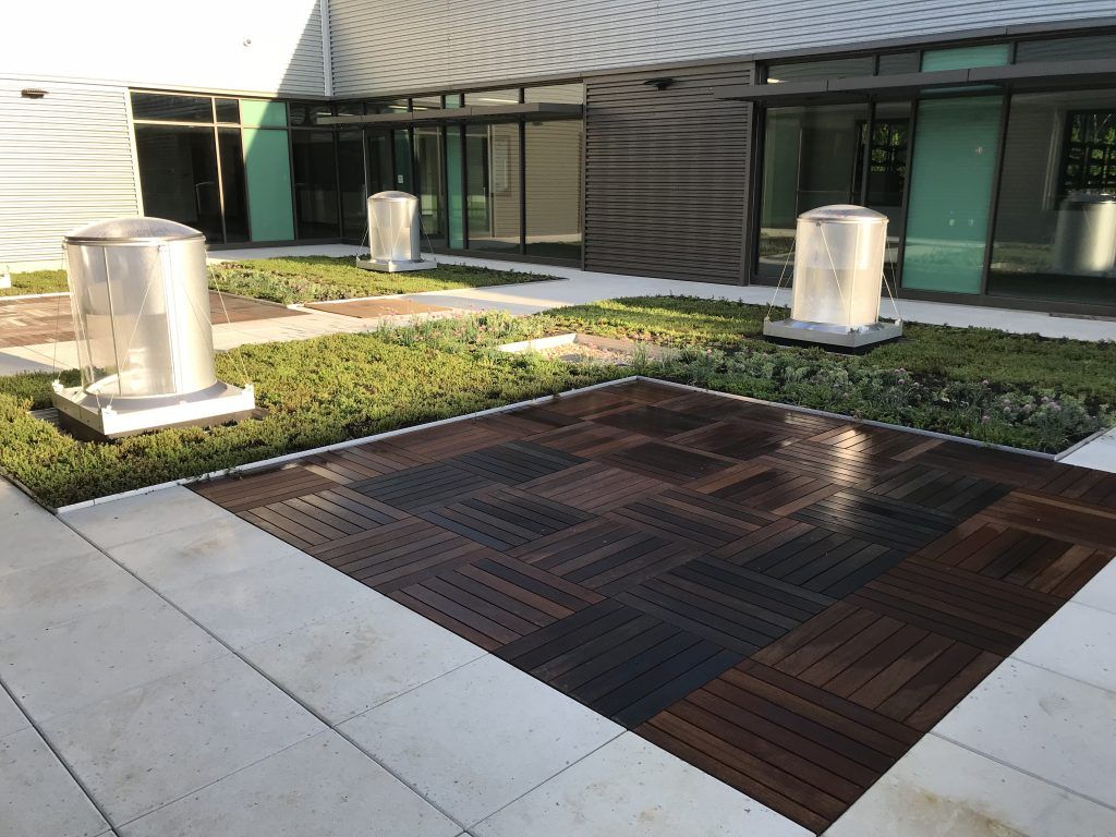 Outdoor patio with dark wood tiles, greenery, and cylindrical objects; building in background.