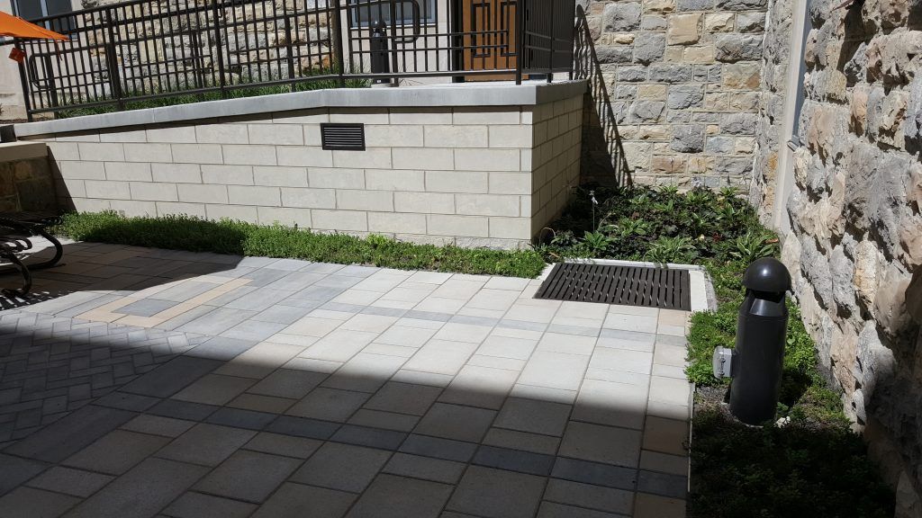 Paved courtyard with a drain, greenery, a brick wall, and a black cylindrical object.