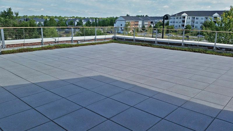 Rooftop patio with gray tiles, metal railings, and a view of apartment buildings and trees under a blue sky.