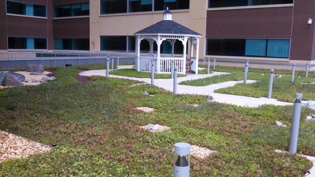 Green roof garden with a gazebo. Beige walkway, tall light posts, and brick building background.