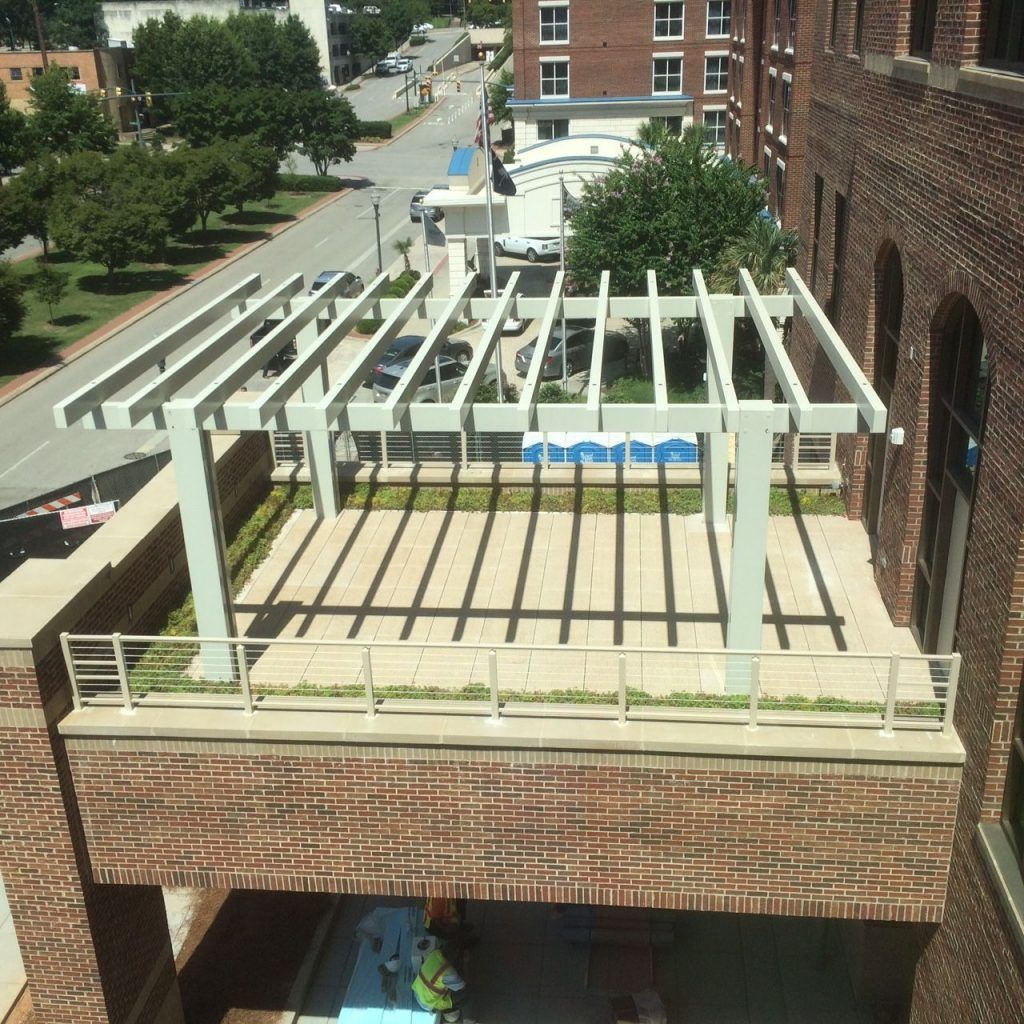 Rooftop patio with pergola casting shadows on concrete. Brick building, street, trees, and sky visible.