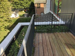 Wooden deck with black railing and hedge border, next to a concrete planter and building.