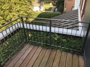 Balcony with wooden floor, black railing, and green foliage; view of trees and building.