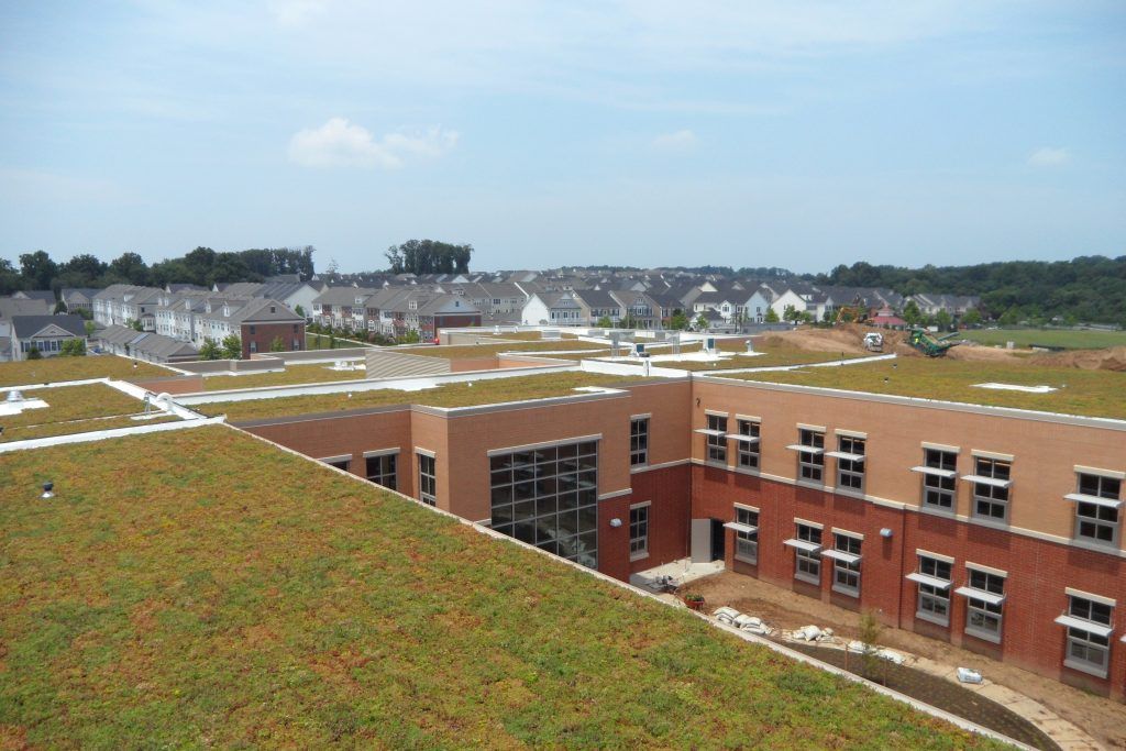 Green roof of a brick building with rows of windows, houses in the background, blue sky.