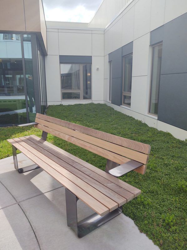 Wooden bench on a concrete patio in a green-covered courtyard next to a building with gray and white walls.