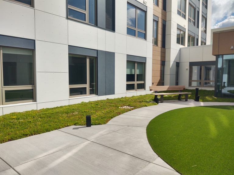 Courtyard with concrete path, green grass, and modern building with windows. A bench sits in the center.