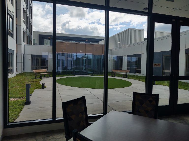 Indoor view through windows of a courtyard with grass, benches, and buildings.