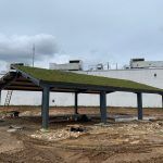 Green-roofed shelter under construction; gray metal supports, dirt ground, white building in the background.