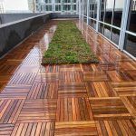 Wooden deck with a green rectangular garden, between a building and a glass wall.
