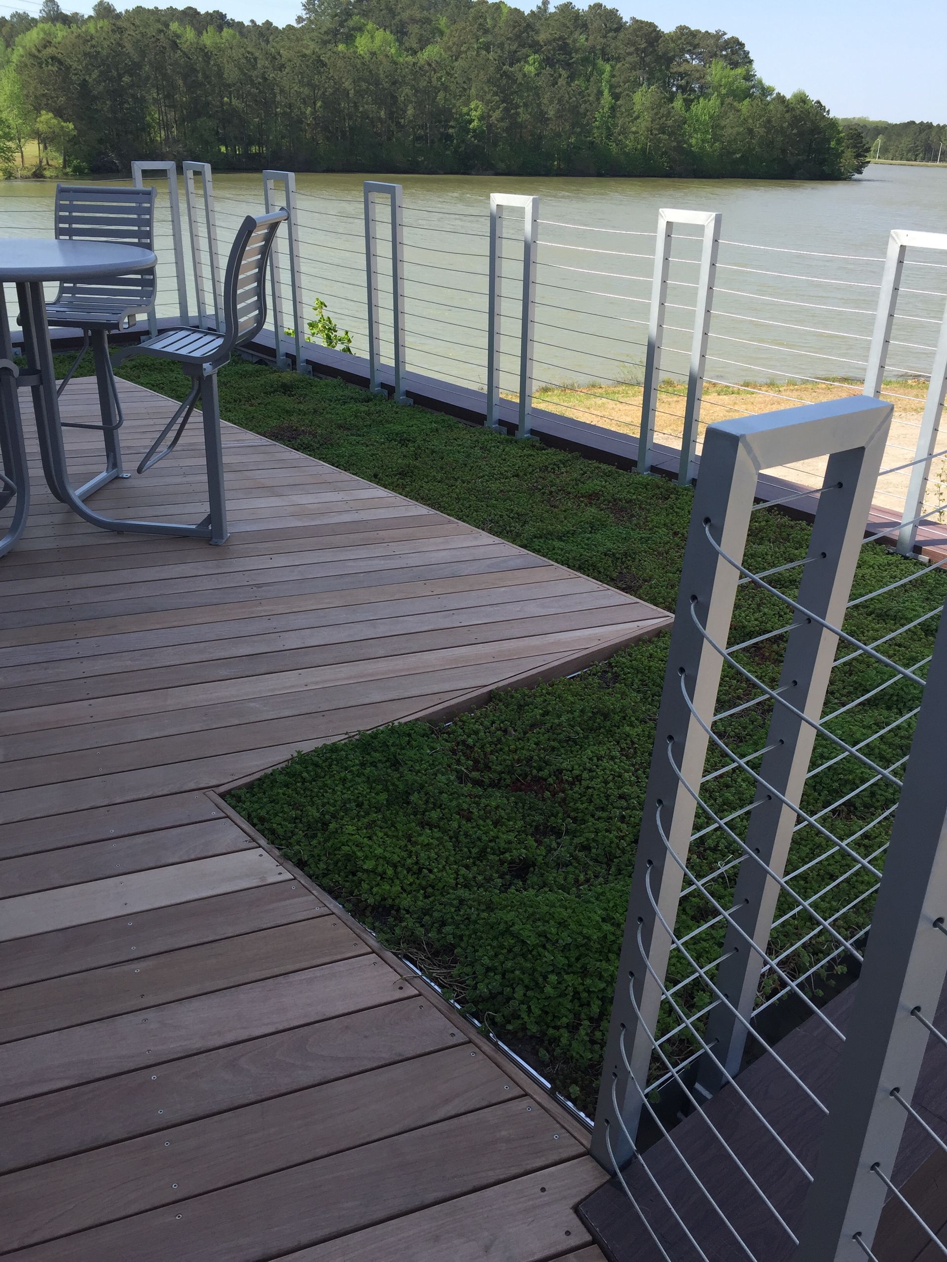 Deck overlooking water with metal railing, table, chair, and green ground cover.