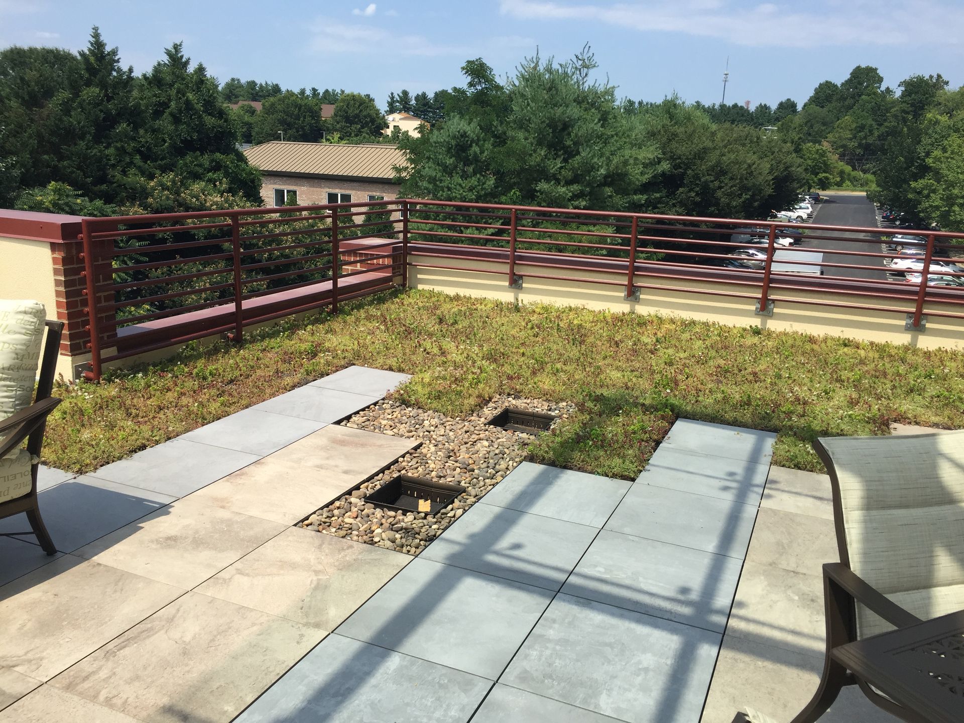 Rooftop patio with green vegetation, gray pavers, and brown metal railing. Sunny day.