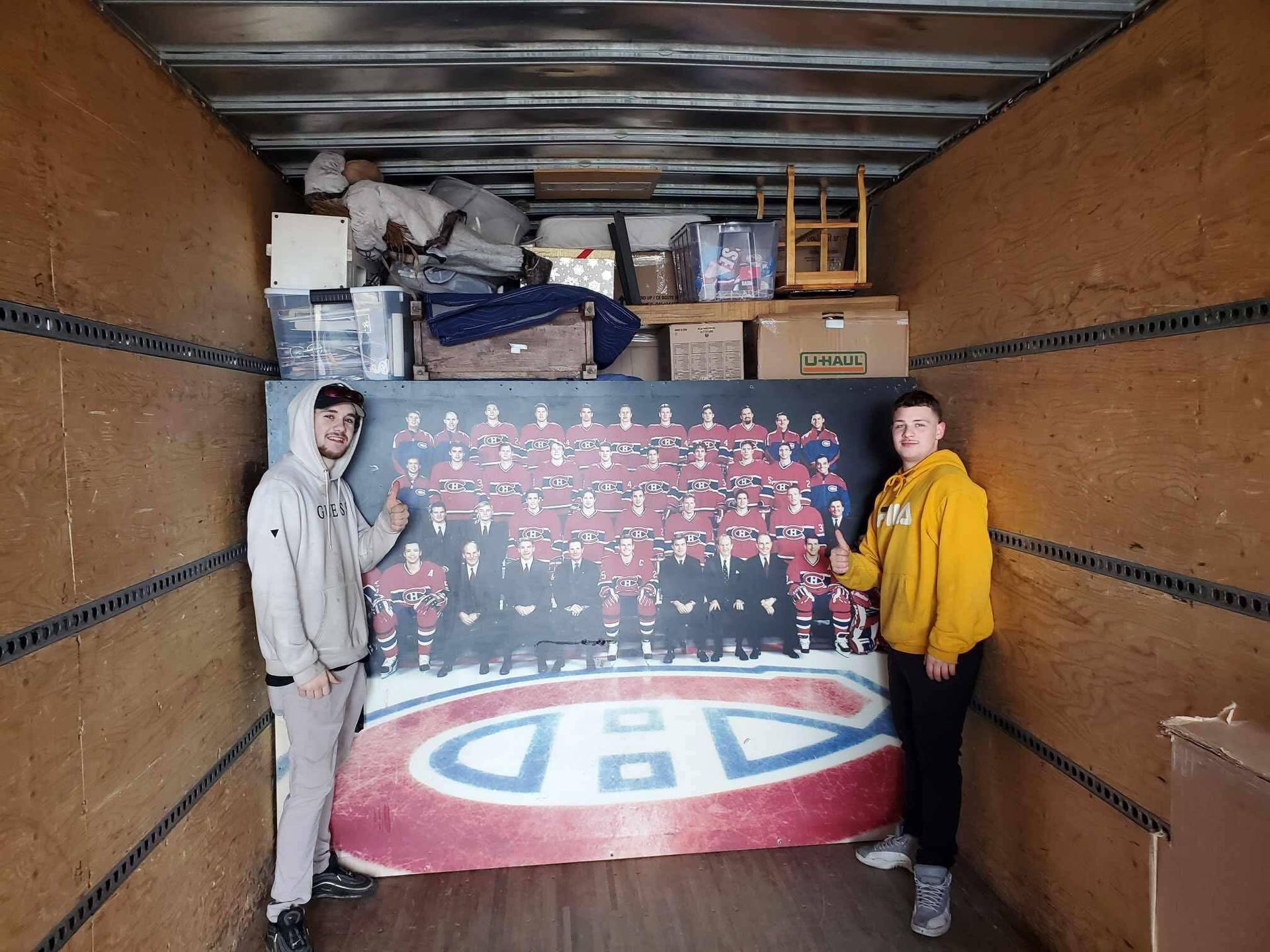 Deux personnes tiennent une grande photo de l'équipe des Canadiens de Montréal dans un camion. Des cartons et des meubles se trouvent également dans le camion.