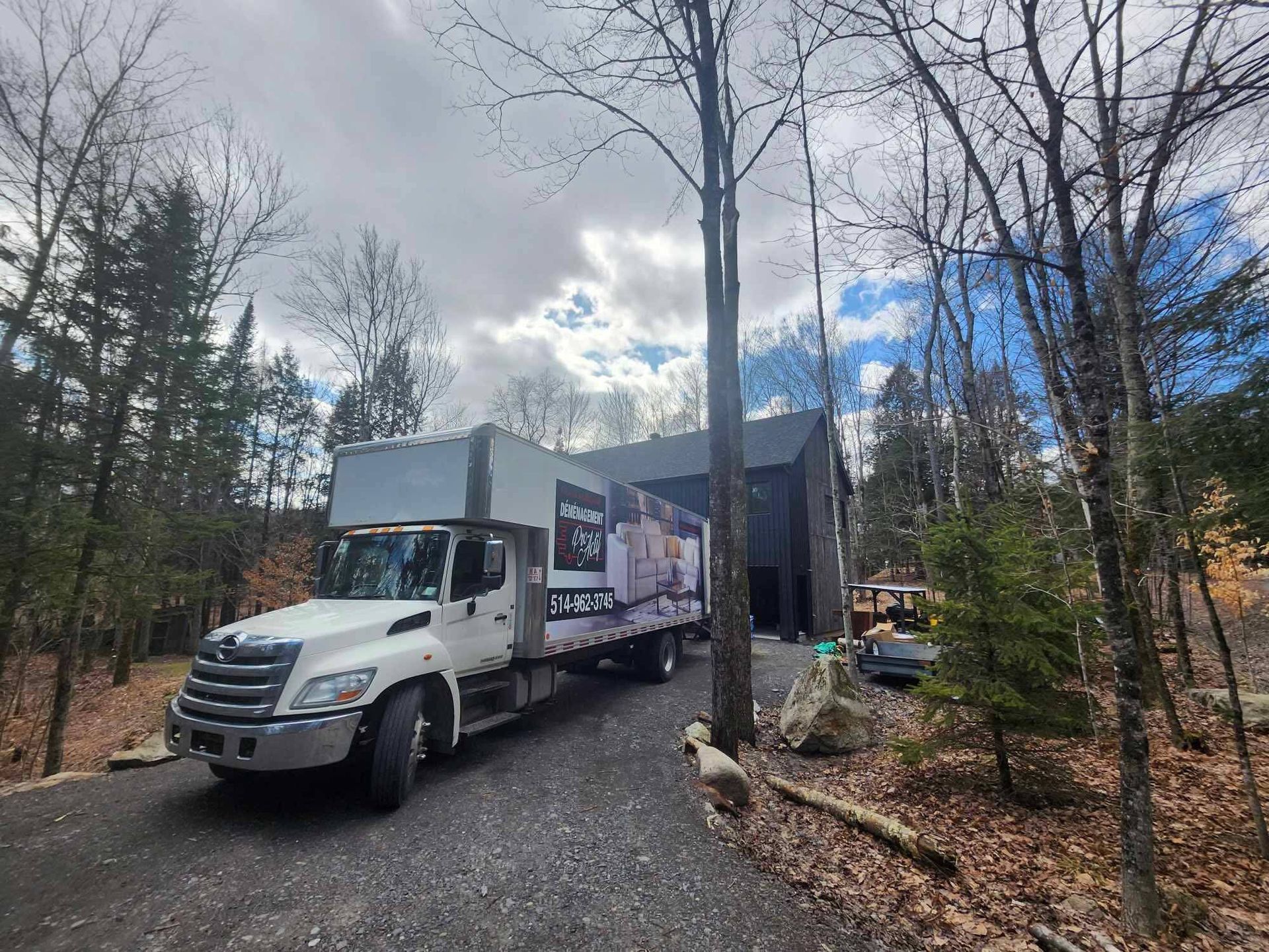Un camion de livraison est stationné sur une allée de gravier près d'une maison de couleur sombre, entourée d'arbres.