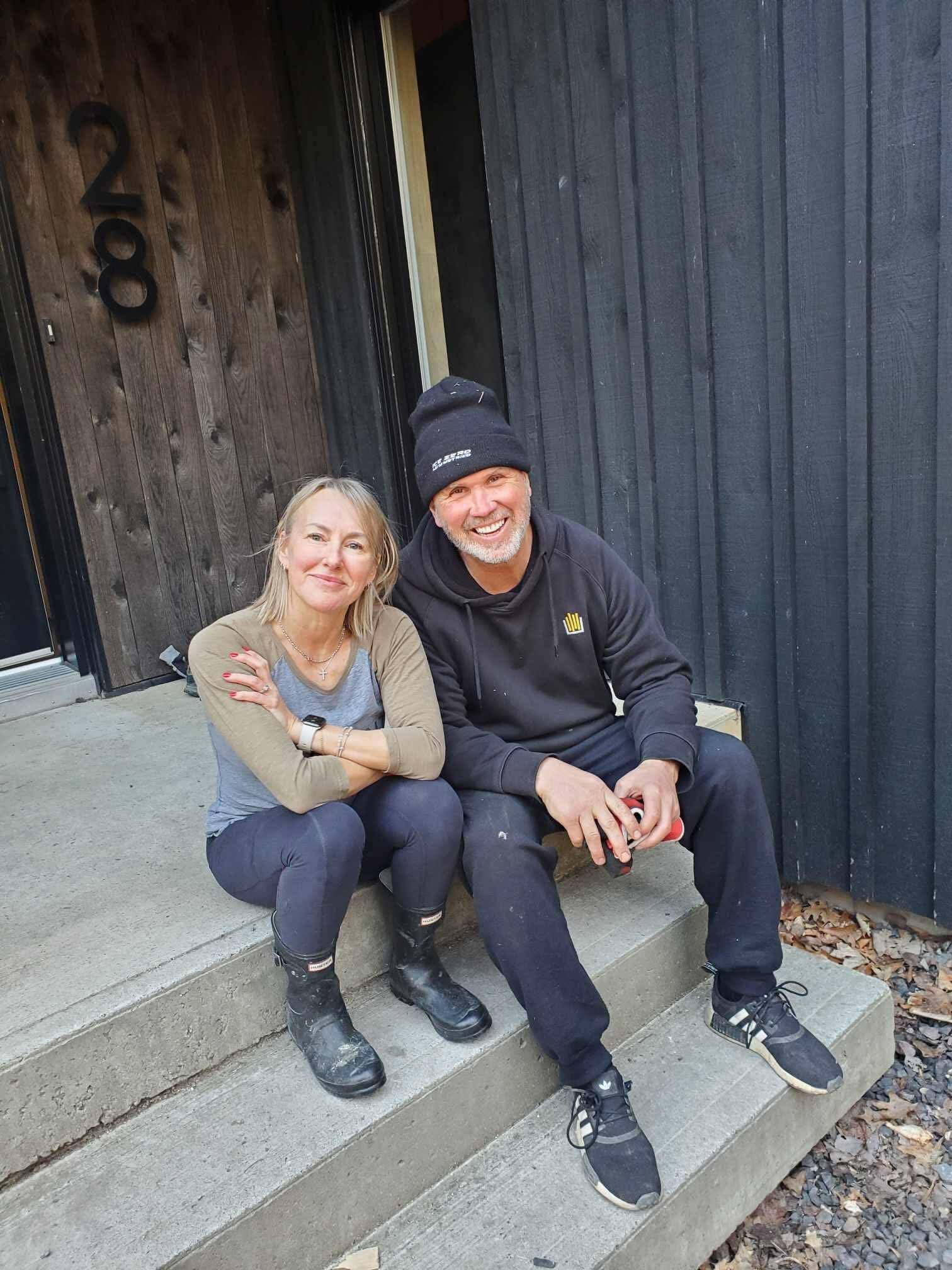 Un couple est assis sur les marches en béton d'une maison noire moderne. La femme porte une chemise claire et des bottes ; l'homme porte un chapeau noir, un sweat à capuche et un pantalon.