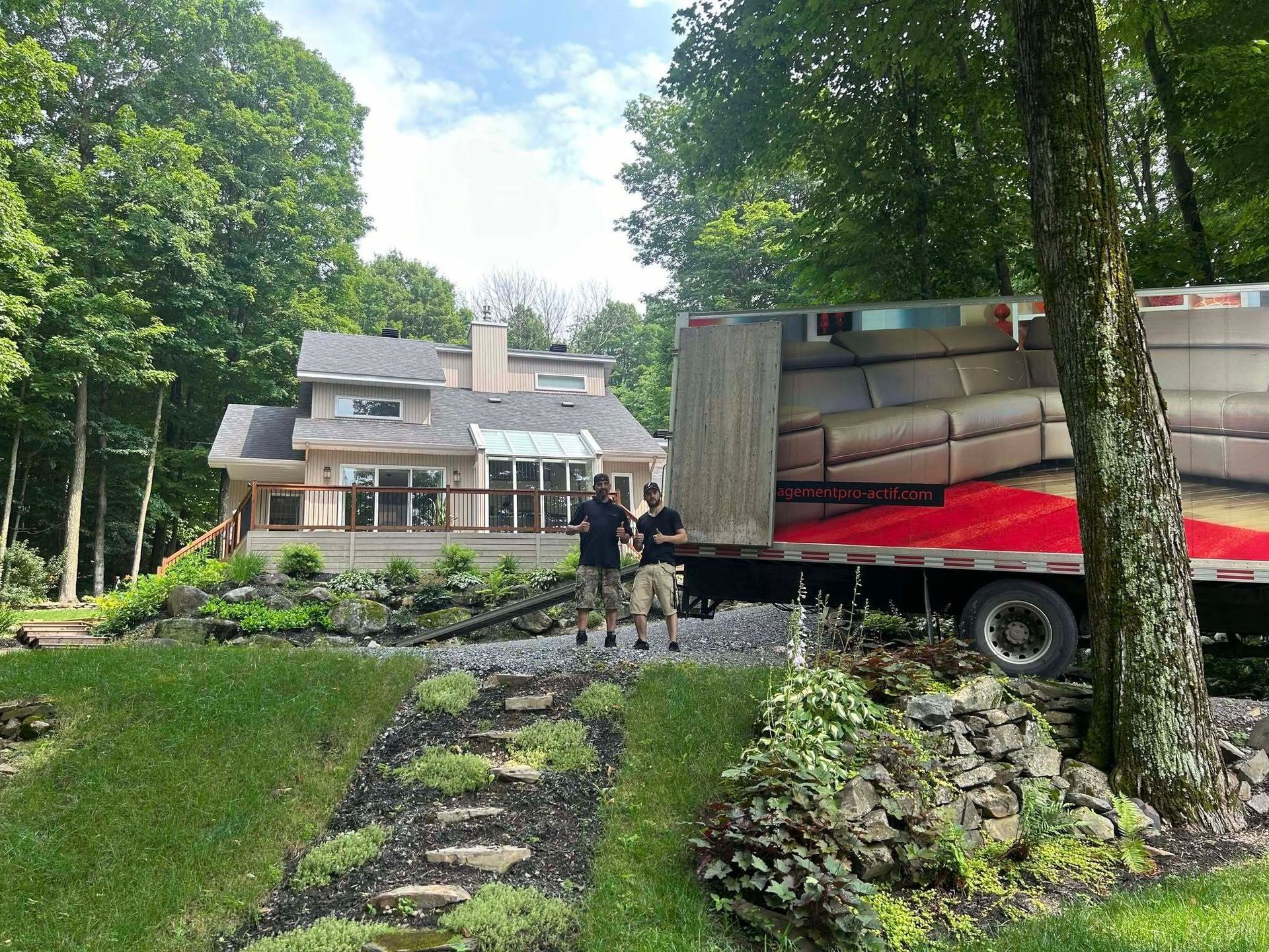 Deux hommes se tiennent près d'un camion de déménagement stationné près d'une maison entourée d'aménagements paysagers et d'arbres.