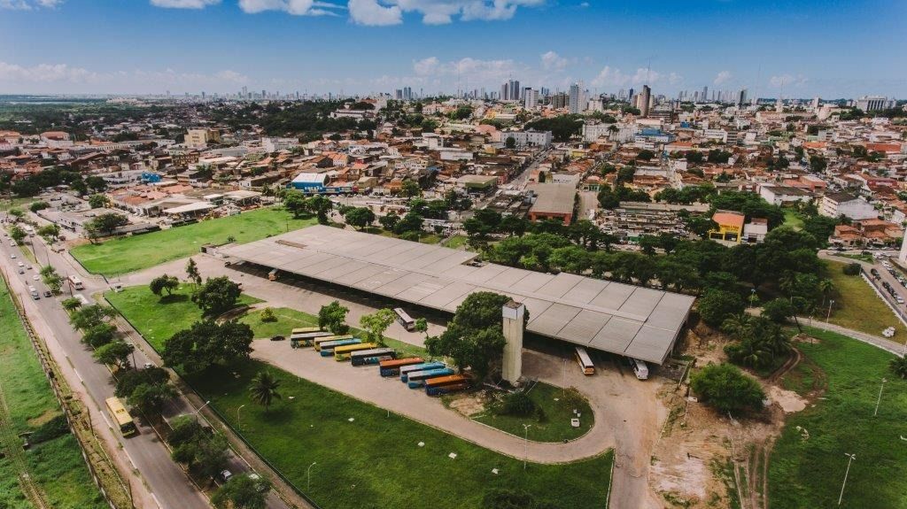 Vista aérea de uma cidade com um terminal de ônibus e expansão urbana ao fundo, sob um céu azul.