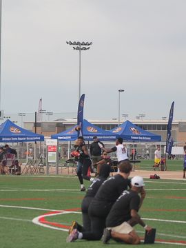 Football game on a green field with players, staff, tents, and sky.