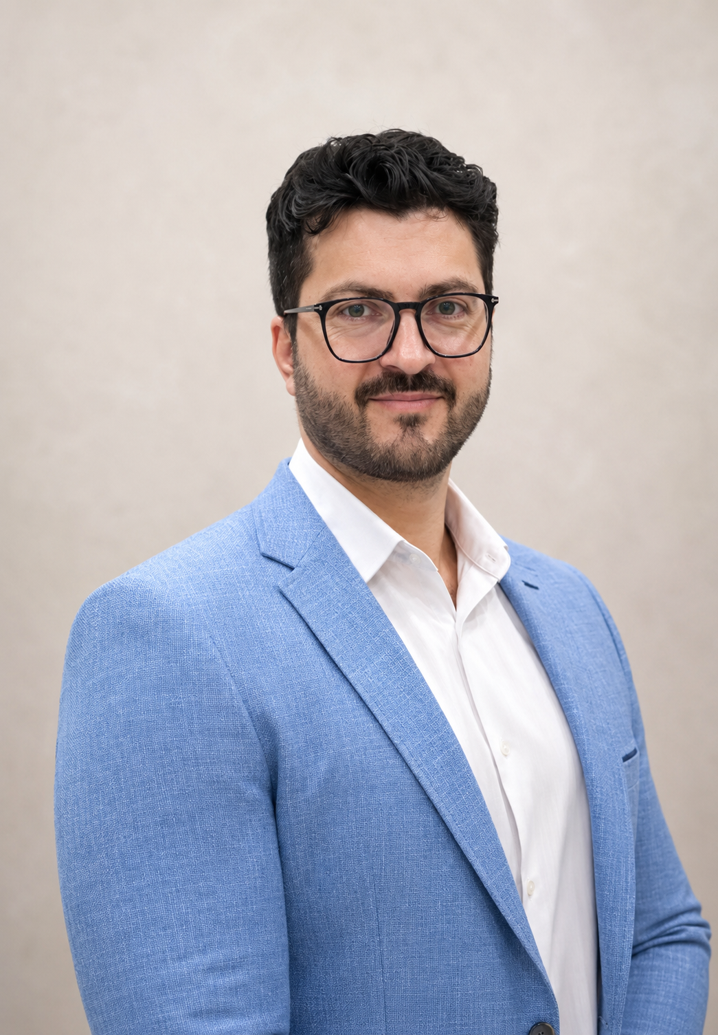 Man in a light blue blazer and white shirt, wearing glasses, posing against a neutral beige background