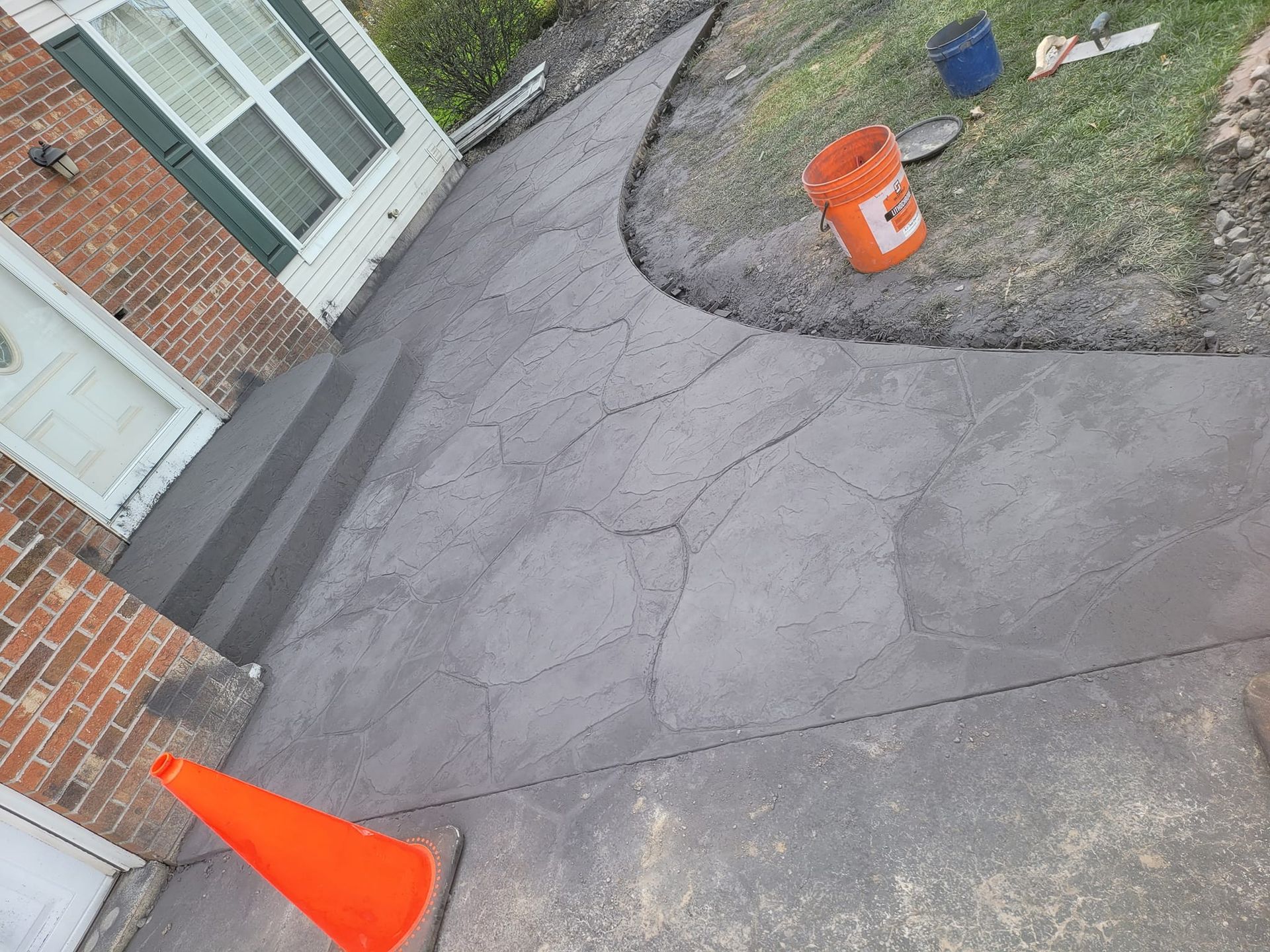 Freshly poured gray concrete walkway with cracks, orange cone and bucket.