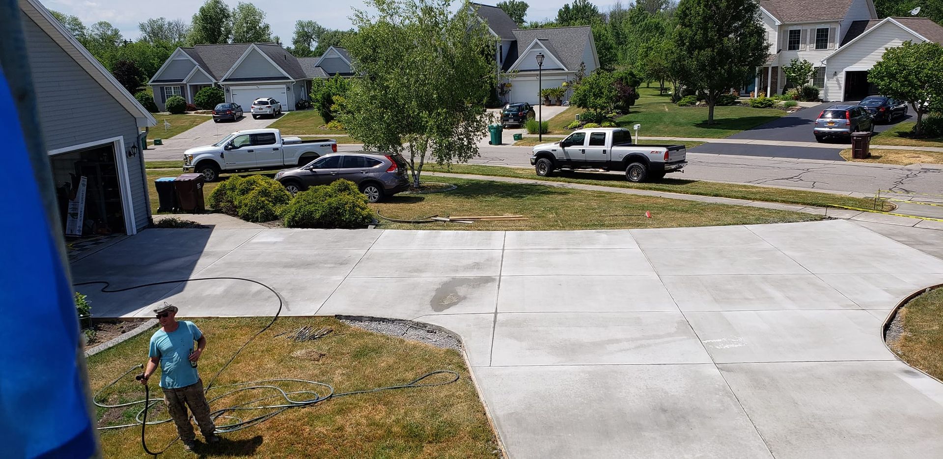 A person waters the lawn in front of a house. Several cars are parked on the street.