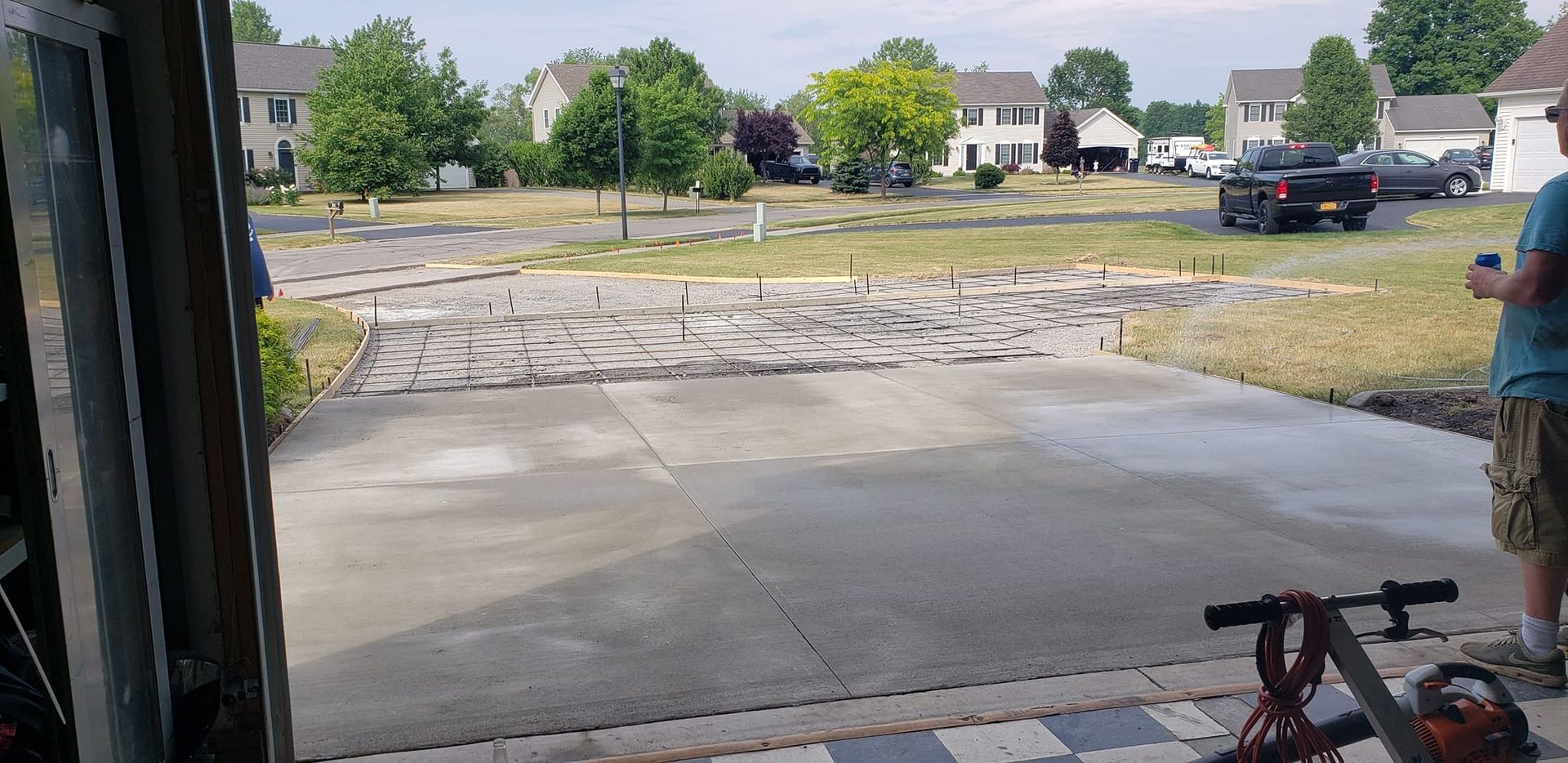 Concrete driveway under construction; houses and trees in background. Person standing on the right with a drink.
