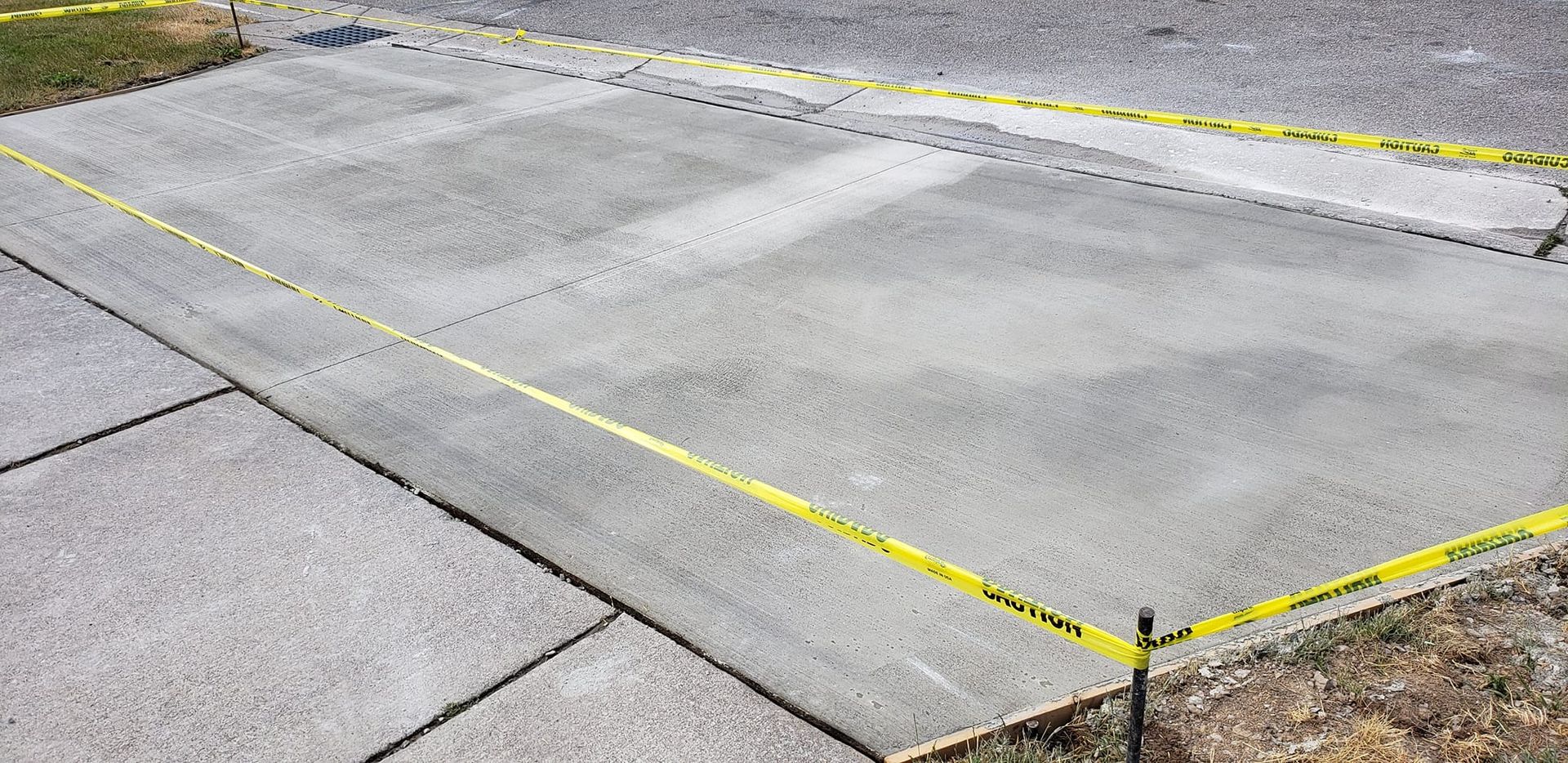 A freshly poured concrete pad is surrounded by caution tape on a paved area.