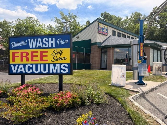 Blue and white car wash building with a large sign reading “FREE Self VACUUMS” and flower bed in front