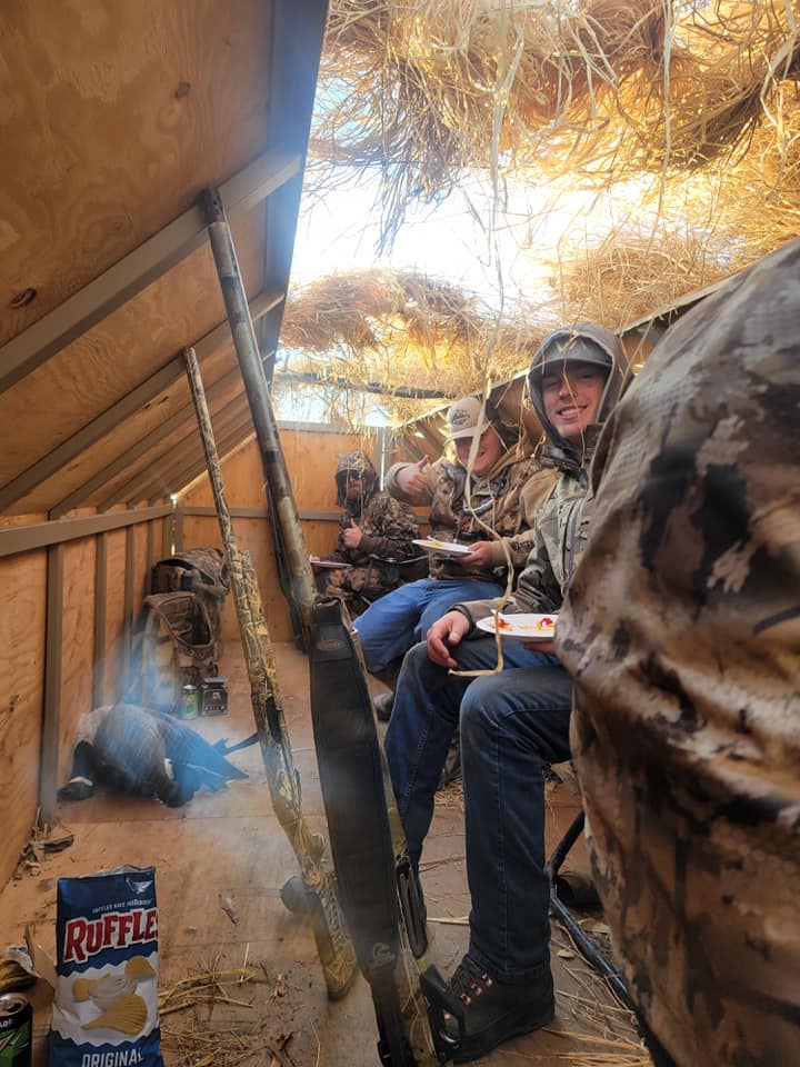 A group of people are sitting in a duck blind eating chips.