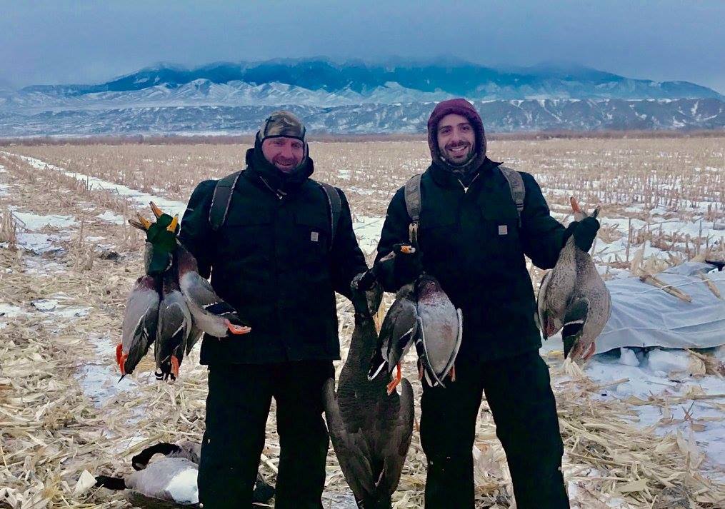 Two men are standing in a field holding ducks in their hands.