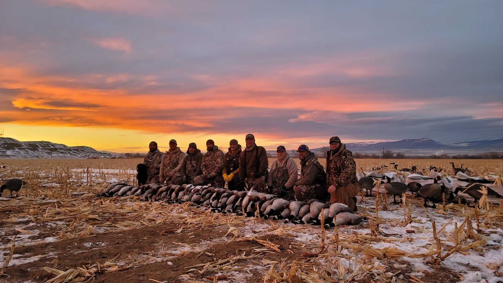 A group of hunters are posing for a picture in a field with a sunset in the background.