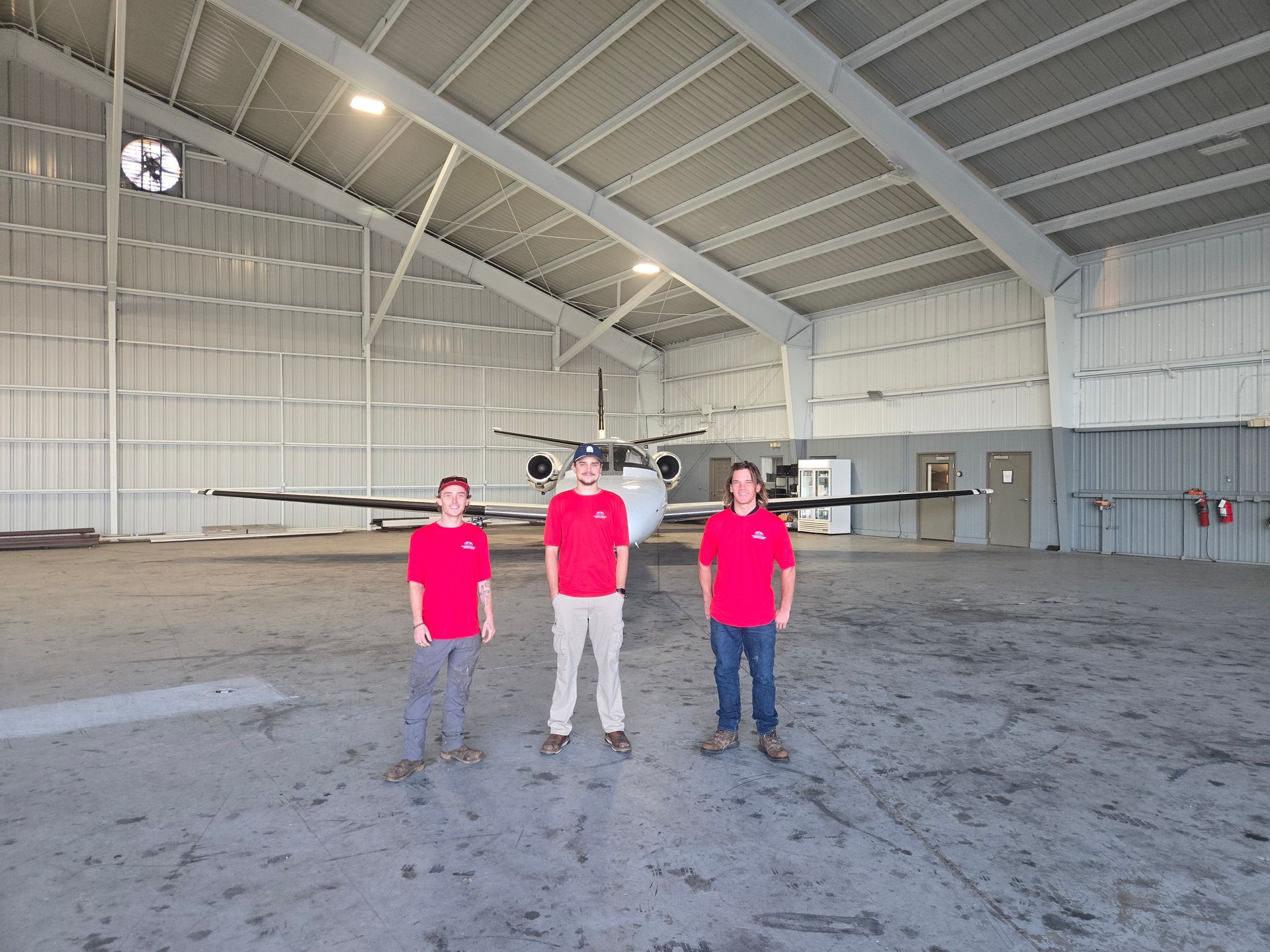 Three people in matching red shirts stand in a large airplane hangar in front of a parked aircraft.