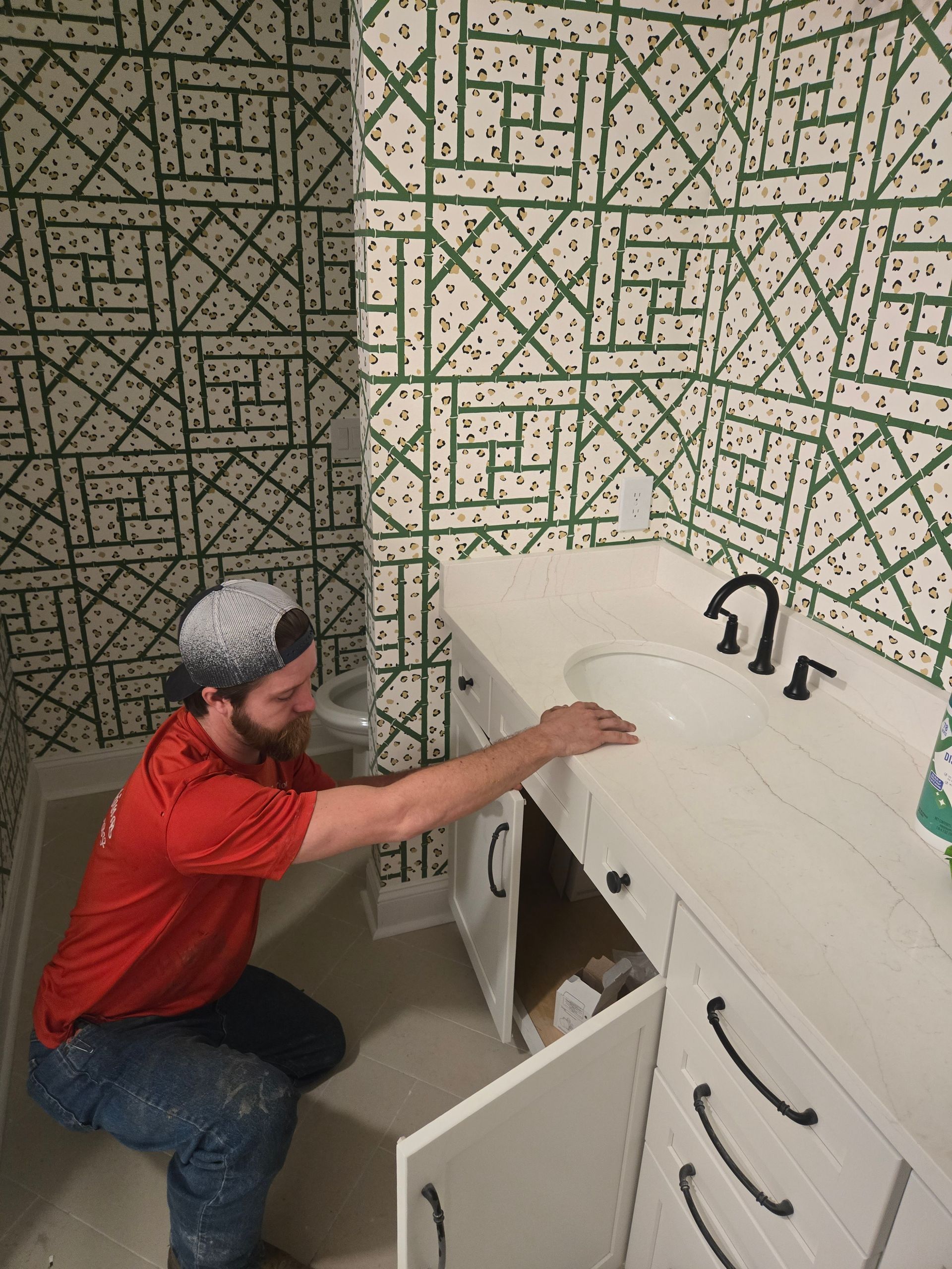 A worker in a red shirt kneels to install or inspect a white bathroom vanity against patterned green and white wallpaper.