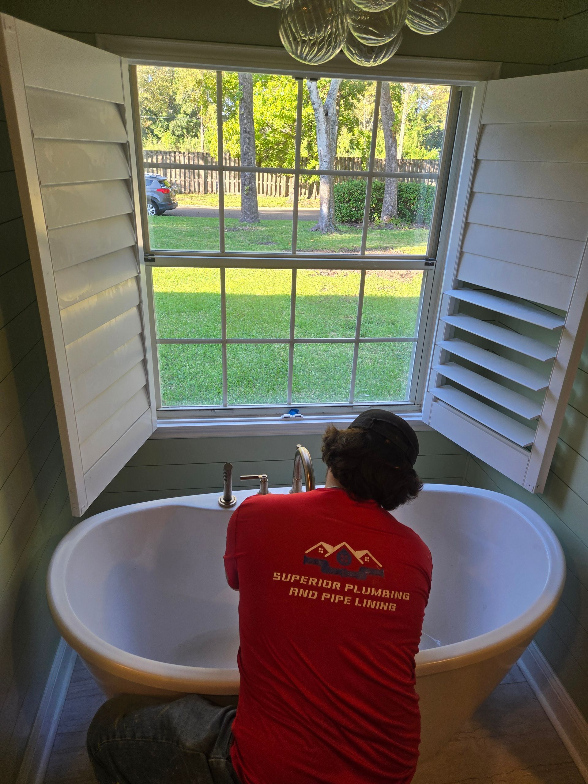 A worker in a red shirt performs maintenance on a white freestanding bathtub positioned in front of a window.