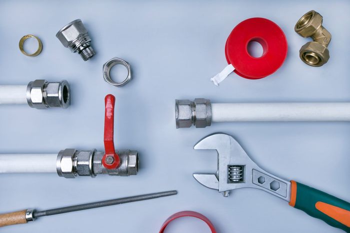 Plumbing tools, metal fittings, white pipes, red thread seal tape, and an adjustable wrench on a light background.