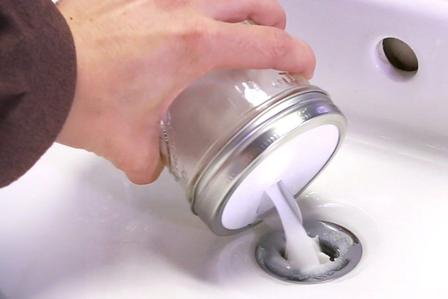 A hand pours a thick, white cleaning paste from a glass mason jar into a white bathroom sink drain.