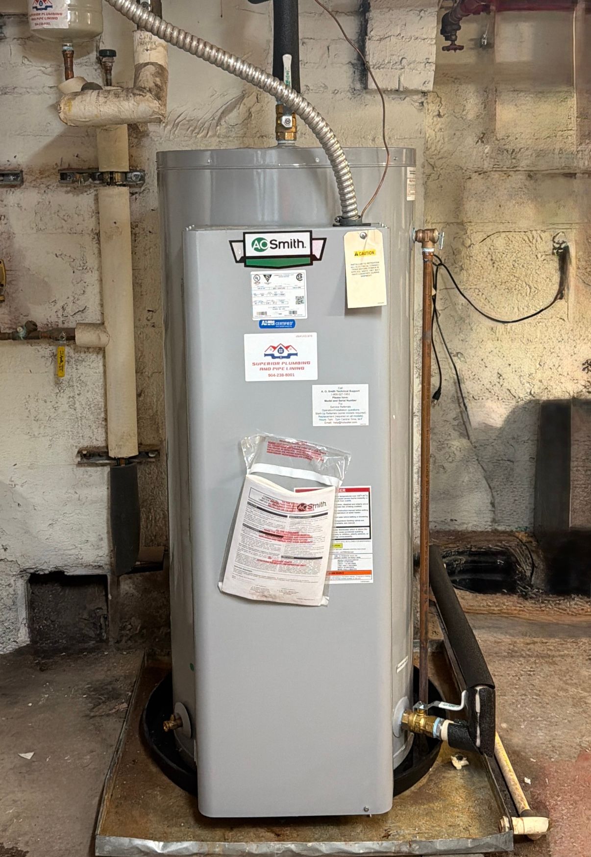 Gray water heater in a basement utility area with pipes, vents, and posted service labels.