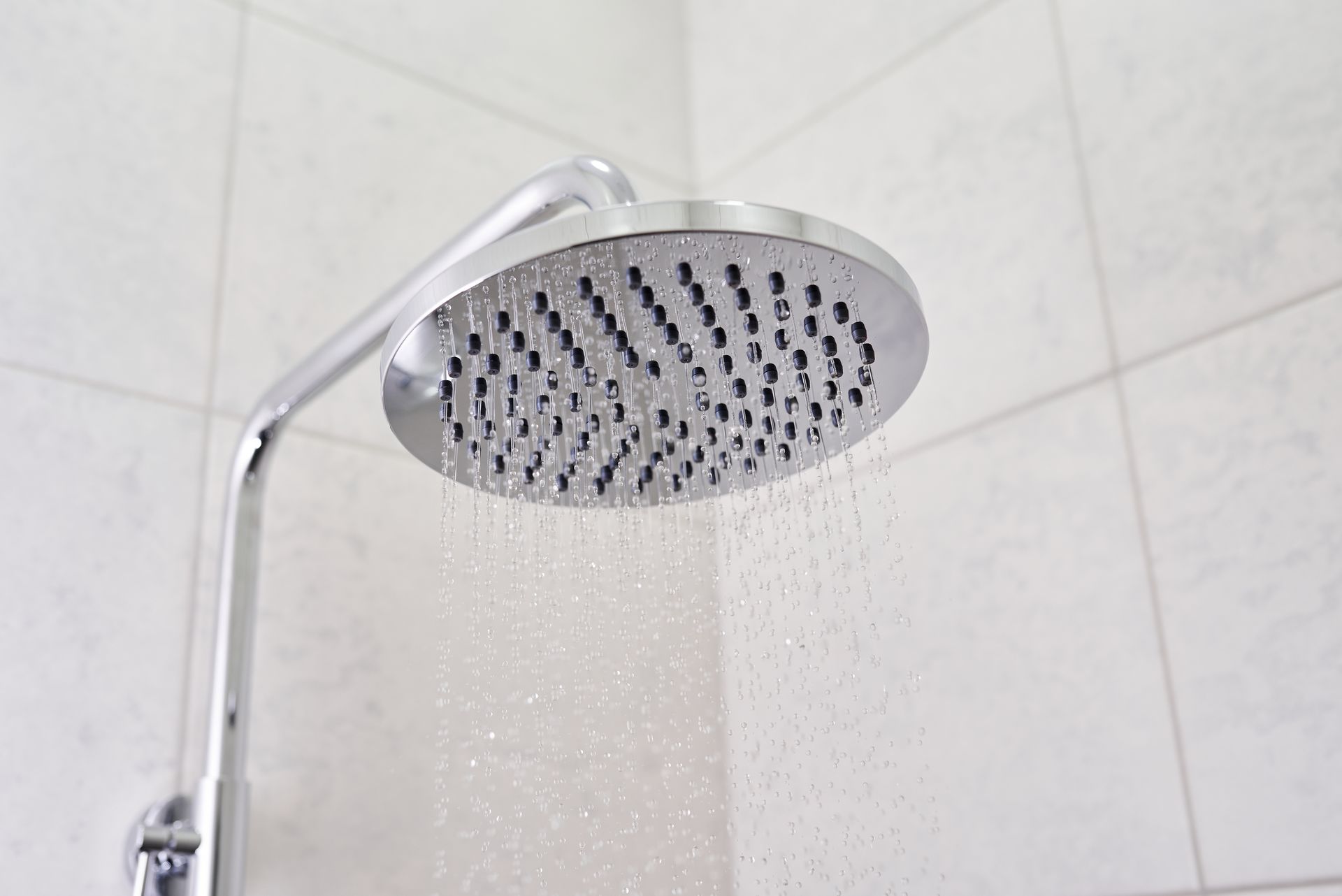 A round shower head mounted on a wall in a tiled bathroom, spraying a steady stream of water downward.