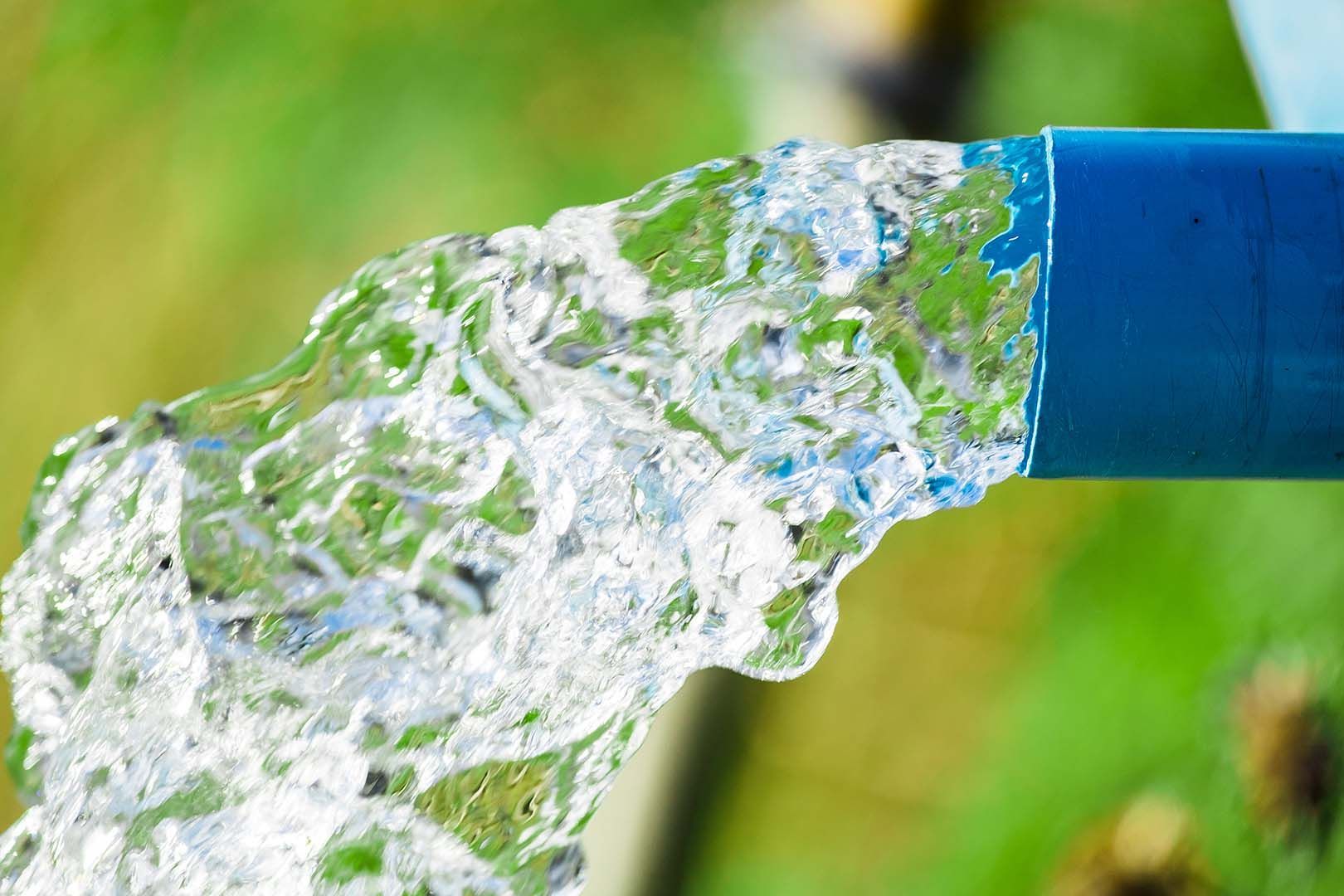 Water flows rapidly from the opening of a blue plastic pipe against a blurred green background.