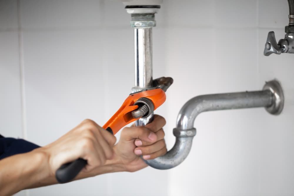 Hands using an orange pipe wrench to tighten or adjust a metal sink drainage pipe under a counter.