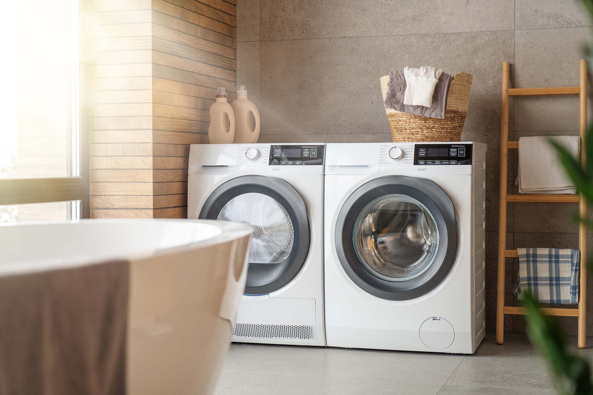 Side-by-side white washer and dryer in a bright, modern laundry room, with a wooden towel ladder and a basket of linens.