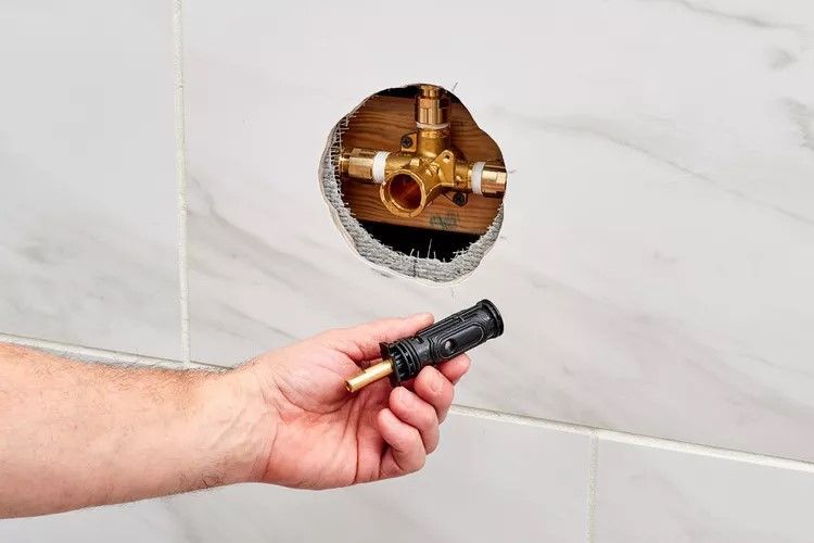 A person holds a black shower valve cartridge in front of an exposed brass plumbing valve installed in a tiled wall.