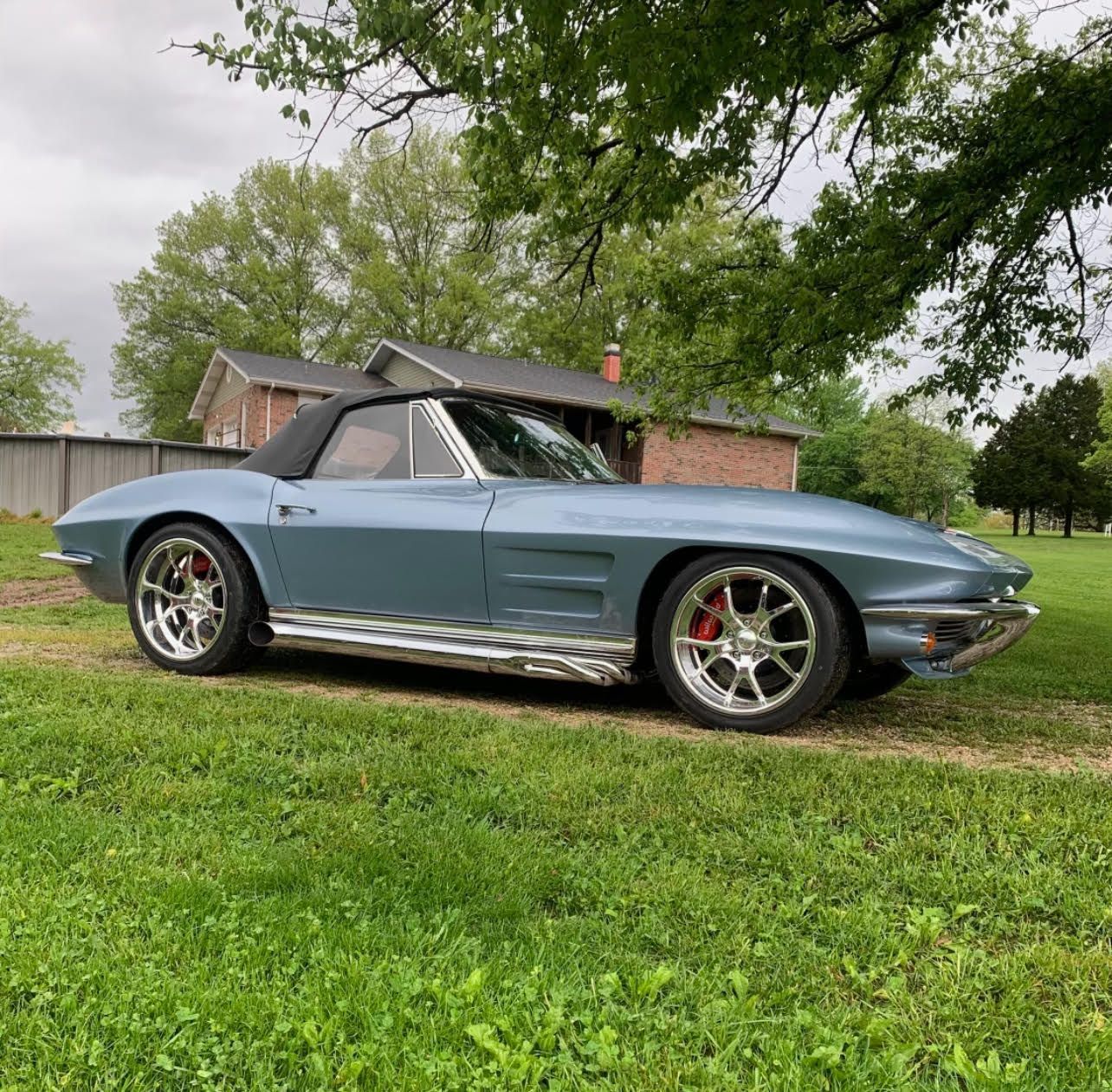 Light blue classic convertible sports car parked on grass near a house and trees.