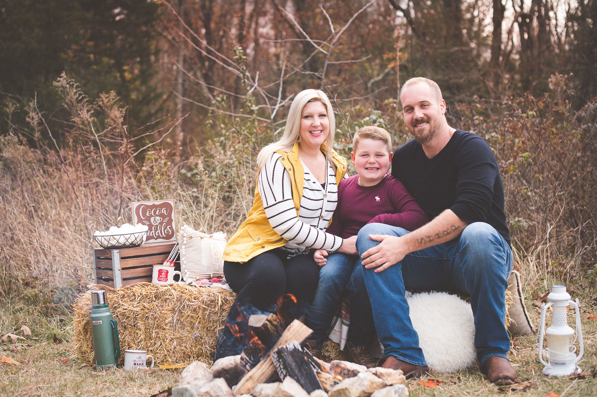 Family seated outdoors by a fire pit in a rustic autumn setting, smiling at the camera.