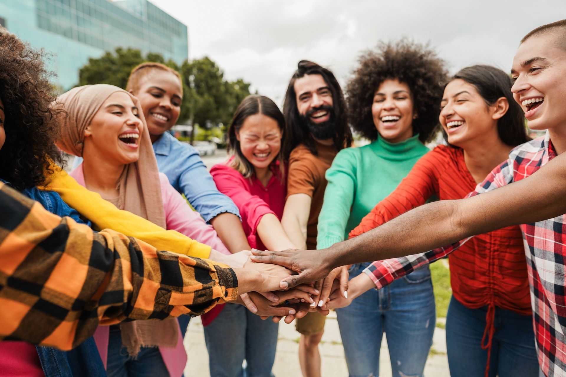 Group of smiling people stacking hands together outdoors in a circle, wearing colorful casual clothes