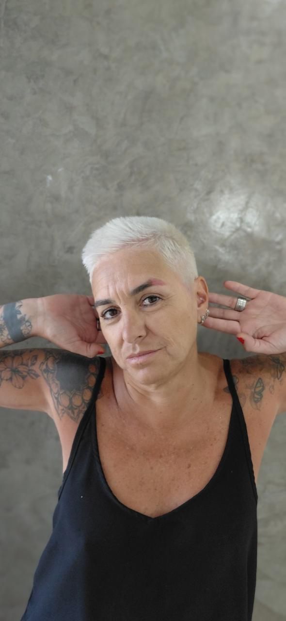 Persona con el pelo corto y blanco y tatuajes, posando con las manos detrás de la cabeza frente a una pared gris.